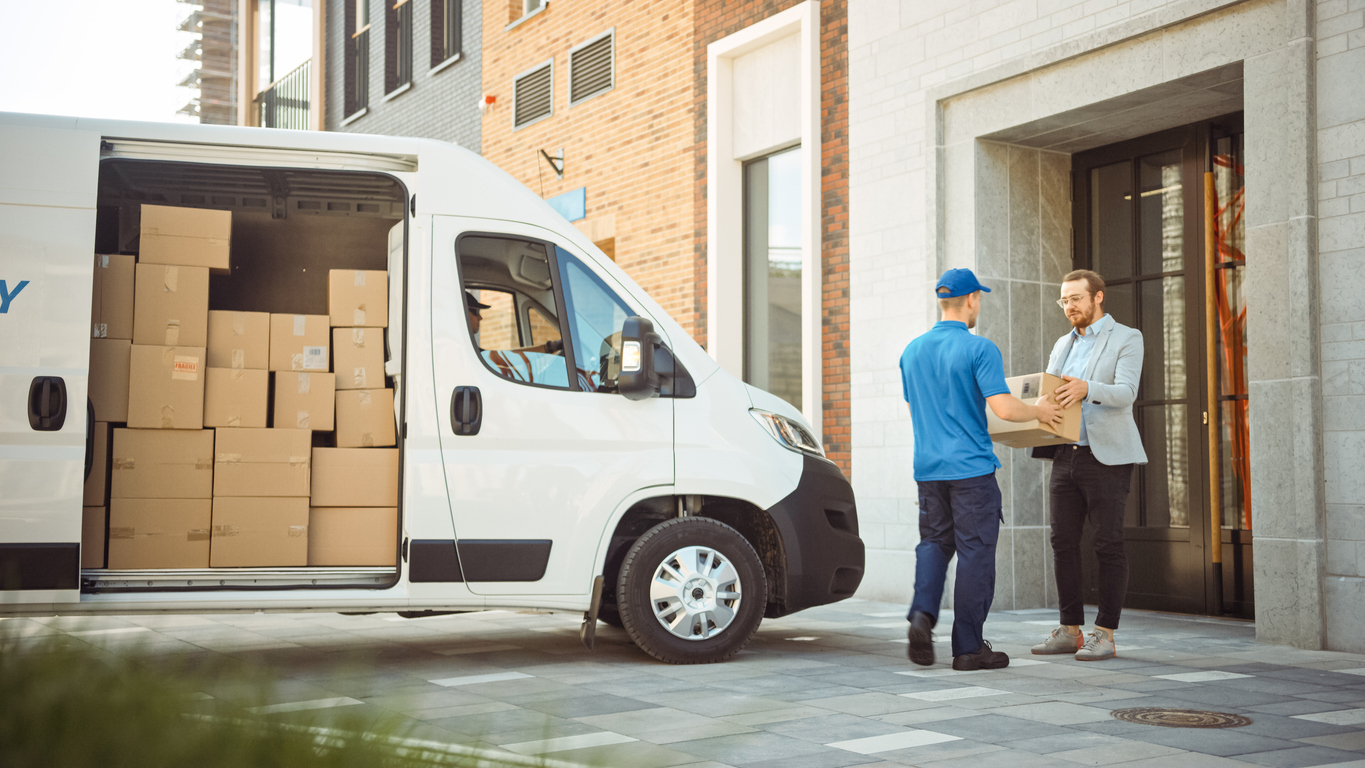 Courier delivers a small parcel cardboard box to a man outside a building. Shipping carrier transports packages from drop-off or pick-up through to the last-mile delivery.